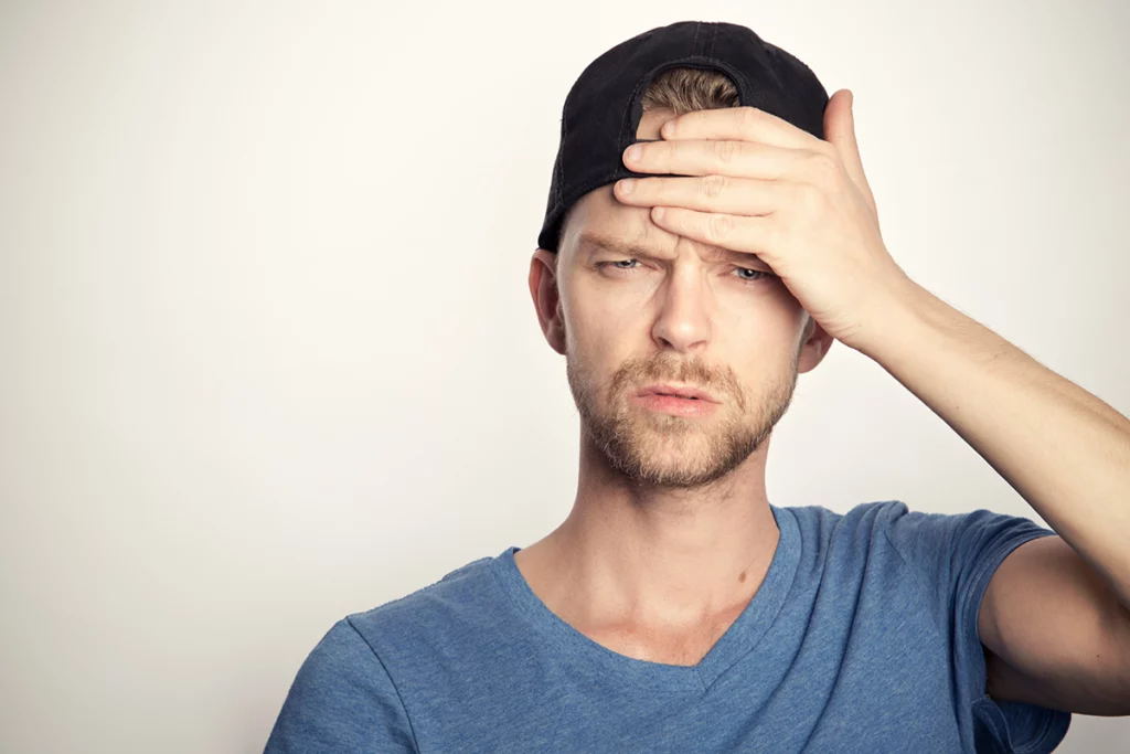 Man in blue t-shirt is shown wearing a black hat backwards while touching his forehead with one hand.