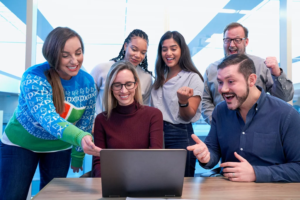 Woman wearing glasses and maroon turtleneck sits at a laptop and smiles. She is surrounded by five people, all of whom are looking at the laptop screen and smiling.