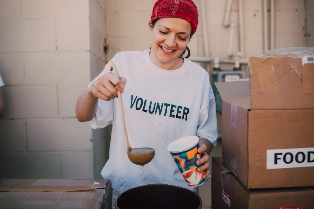 Woman does a small act of care for her community. While wearing a white “volunteer” shirt, she hands out cups of soup.