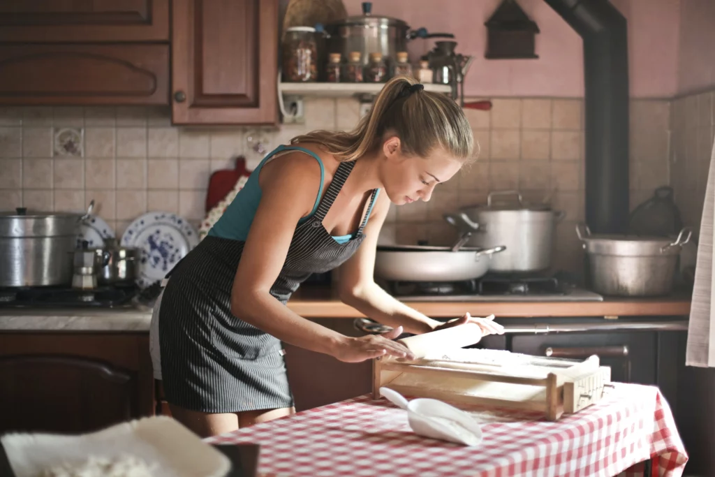 Woman in apron bakes in kitchen on top of a checkered tablecloth. She is preparing her meals for the week.