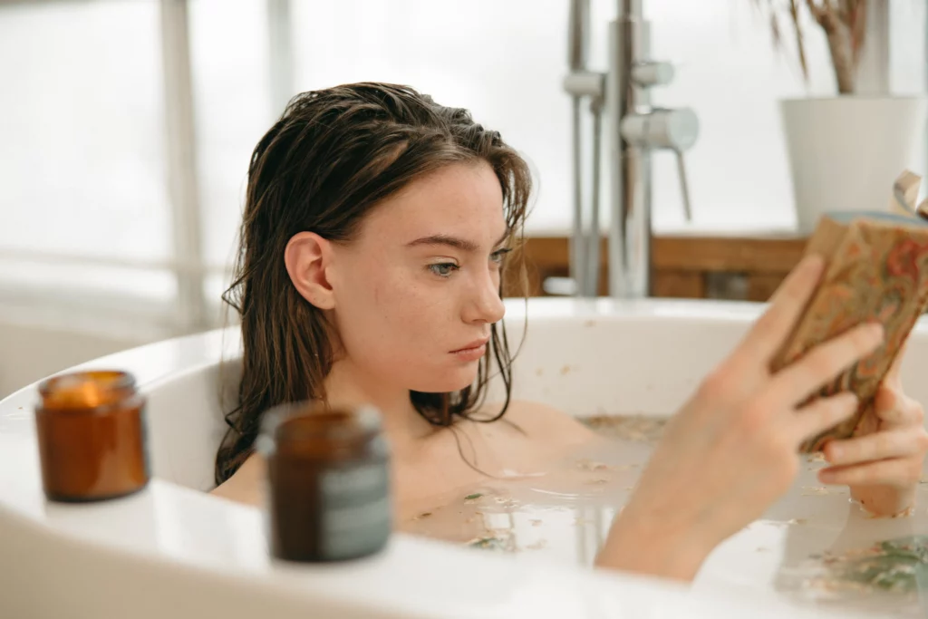 Woman reading book in bathtub to practice self-care. She has candles lit around her.