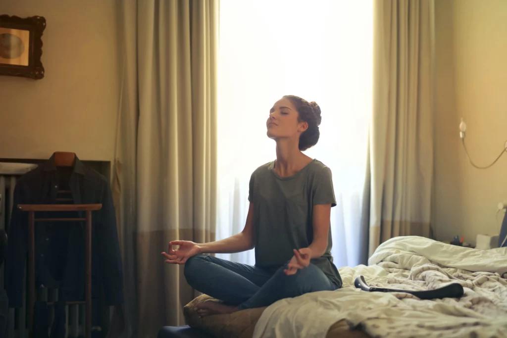 Woman sits on corner of bed in Burmese meditation stance. She slows her breathing to stay calm and find peace.