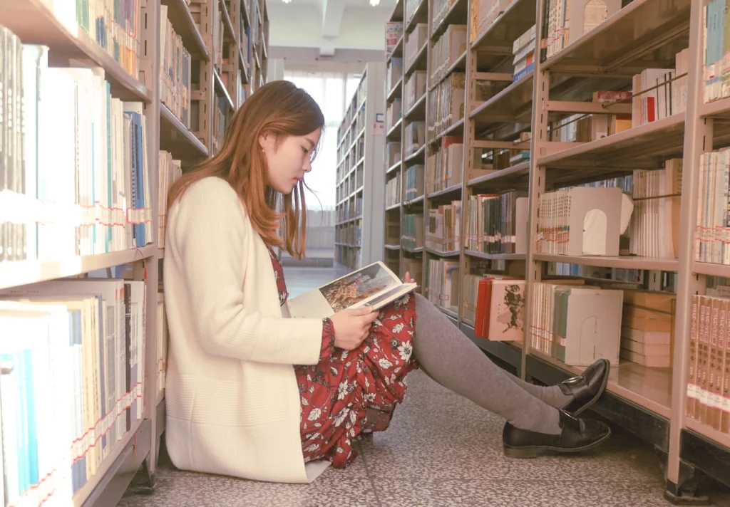 Woman sits down in the middle of a library reading a book.