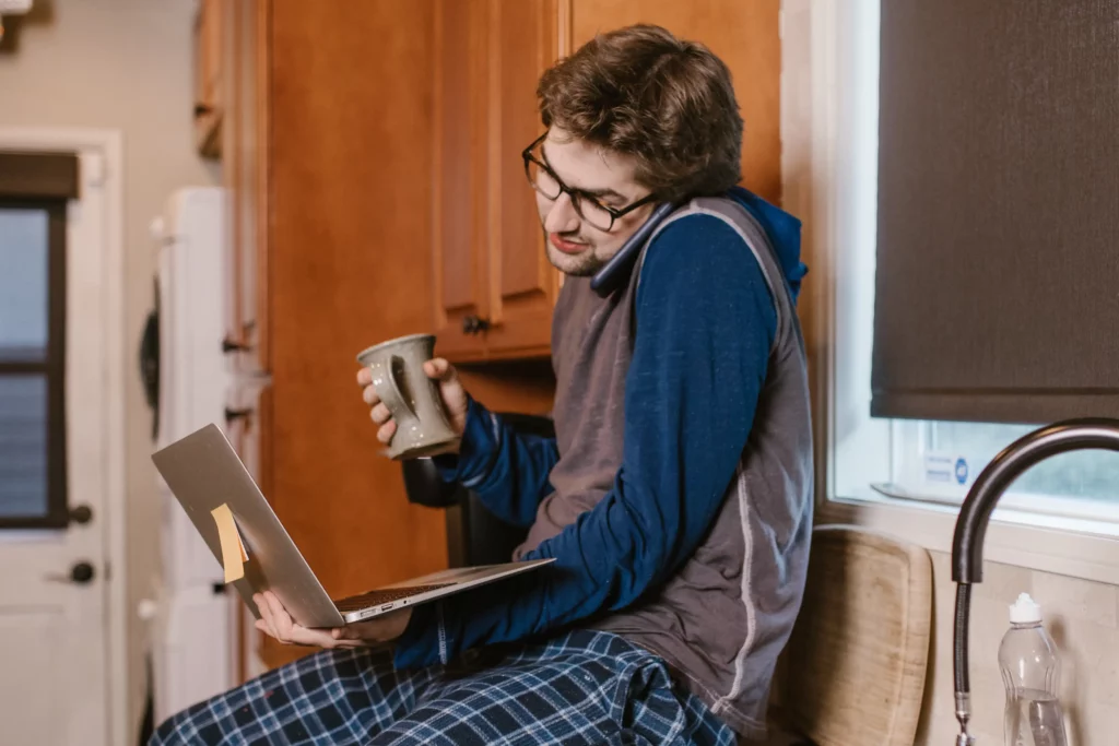 Man sitting on kitchen counter, holding a mug of coffee an a laptop. He’s in pajamas and on the phone talking to his utility provider.