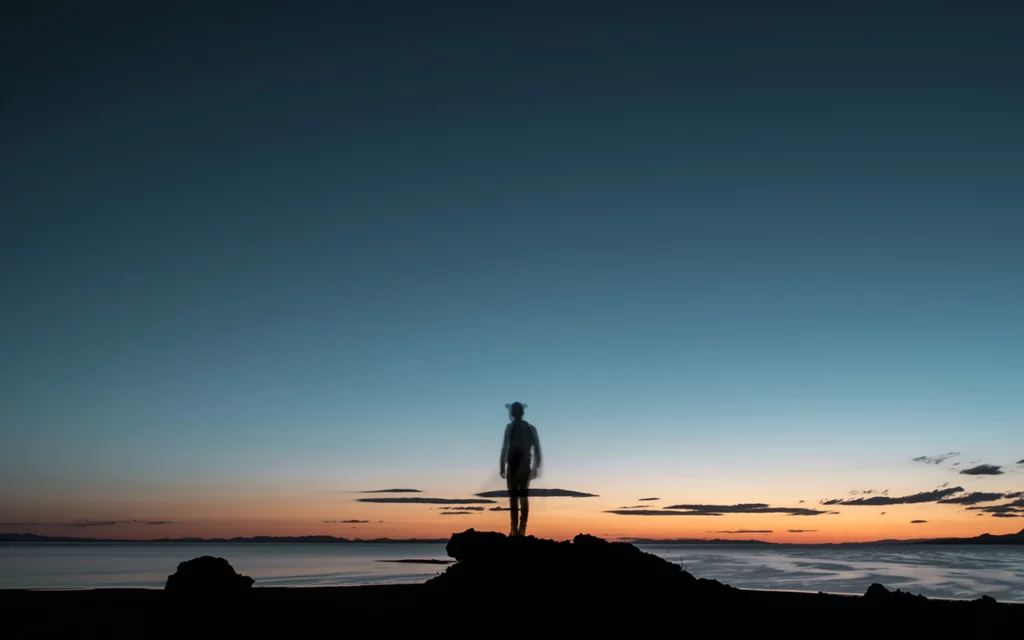 A person stands on a rock on Antelope Island in Utah while the sun sets in the distance.