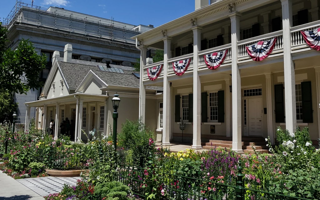 The Beehive House in Salt Lake City has a beautiful porch with columns. It has a lot of history to it. There are flowers growing in the front yard.