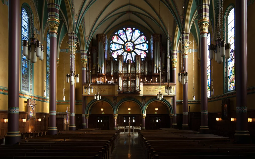 Inside of the gorgeous Cathedral of Madeline. The large pipes of a pipe organ are on display in the front of the church. The center has a bright stained-glass window. Tall, narrow stained-glass windows can be seen on both sides of the building. There are tall columns within the room, helping support the ceiling.