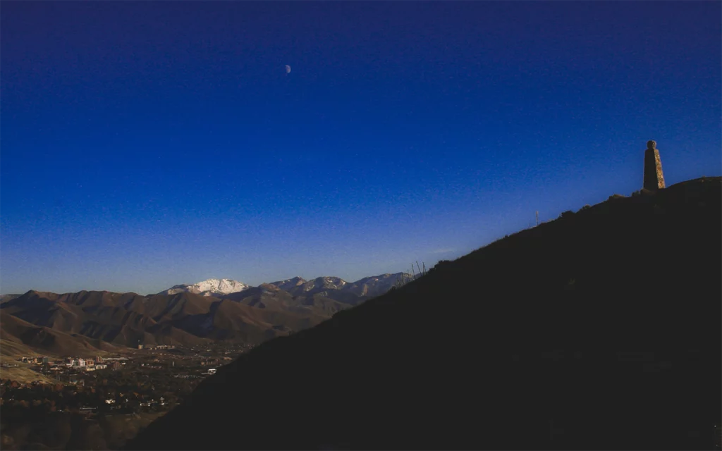 At the top of a hill you see a tall, towering landmark. The landmark is Ensign Peak. From Ensign Peak, you can look down at Salt Lake City, which is in the background of the image. The sun is going down. The sky and moon are clear to see.