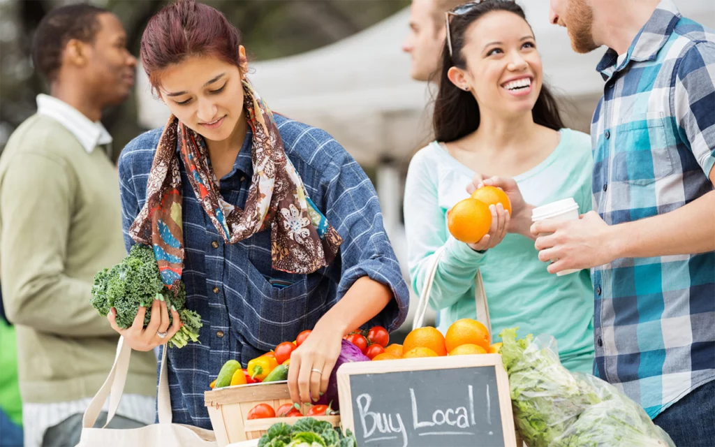 Shoppers look at fresh fruits and vegetables at a farmers’ market. They are looking at tomatoes, cabbage, and oranges. A sign in chalk says, “Buy local!”