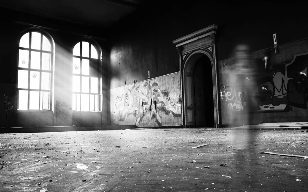 Empty room with debris on the floor and graffiti on the walls. One piece of graffiti says “help”. Sun comes in through the windows of the wall. The room looks abandoned and haunted.