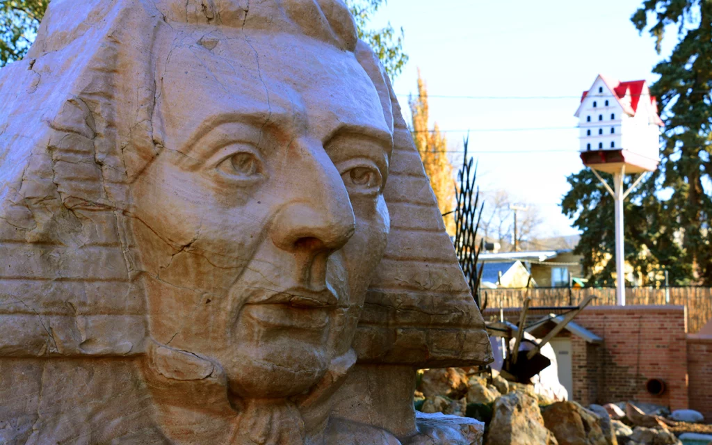 A stone carving sculpture that represents a crouched Sphinx and the face of LDS Church founder Joseph Smith. Behind the sculpture, other unusual and original art pieces can be seen within the Gilgal Sculpture Garden in Salt Lake City