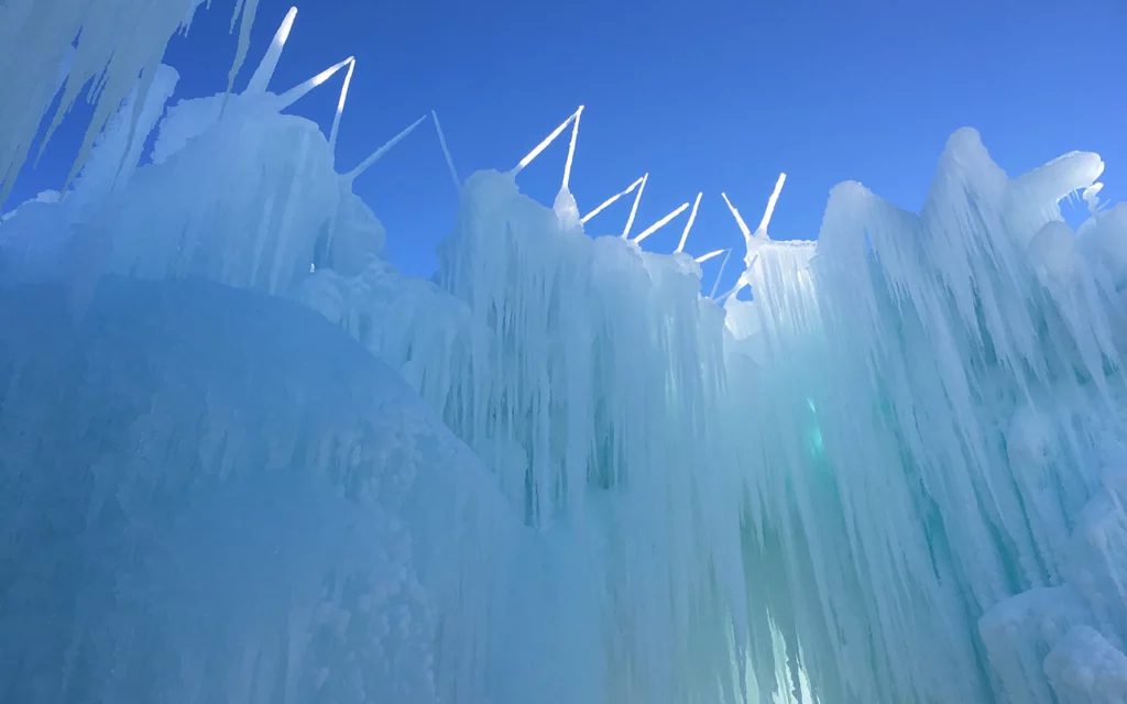 The famous Ice Castle sculptures outside of Salt Lake City. Spiky ice formations adorn the top of the castle. Above it is a blue sky. Sunlight can only reach the top of the castle.