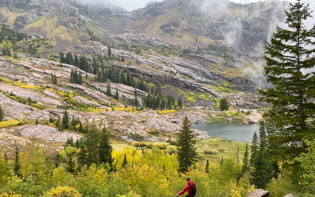 A man from up high overlooks Lake Blanche in Big Cottonwood Canyon. The lake is past many trees. On the other side of the lake are more mountains. Fog is in the air.