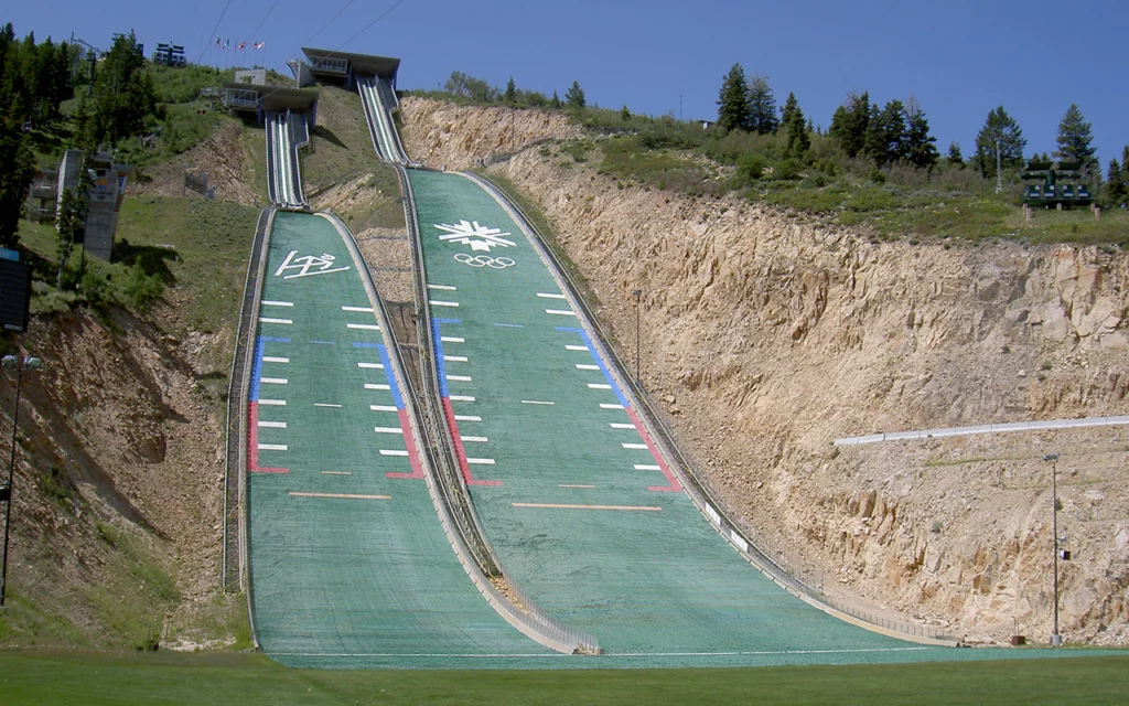 On the side of a tall hill are two steep green slides. One has the image of a person ski jumping. The other slide has the Olympic logo. These slides are located at the Utah Olympic Park, near Salt Lake City, and are used for summer-time ski jumping. The green surface is made from plastic and is feels similar to skiing on snow.