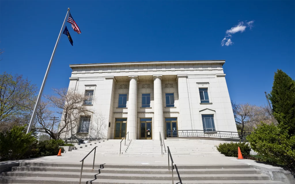 From the outside, the Pioneer Memorial Museum, in Salt Lake City has two floors. This white square building has two columns in the front of it. There are several steps leading up to the entrance. The US flag and Utah flag are waving on a flagpole out front of the building.