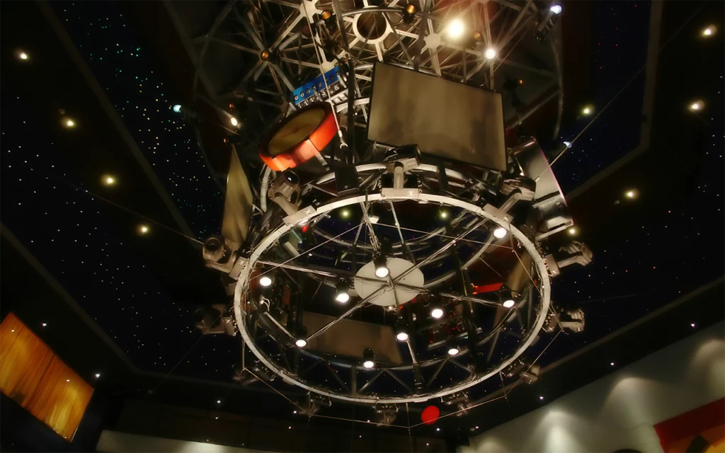A cool futuristic display on the ceiling. It appears to have lights, a clock, and several TV monitors. Above the display is a ceiling made to look like space and stars. This is inside of a planetarium.