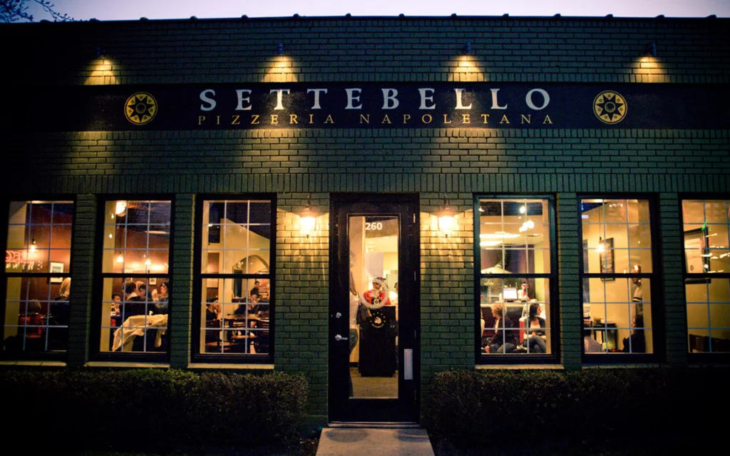 The outside of the Settebello Pizzeria Napoletana restaurant in Salt Lake City. Inside, there is warm light and many people eating food. The host can be seen standing at the podium just past the glass front door. The restaurant is not very big but looks very cozy.