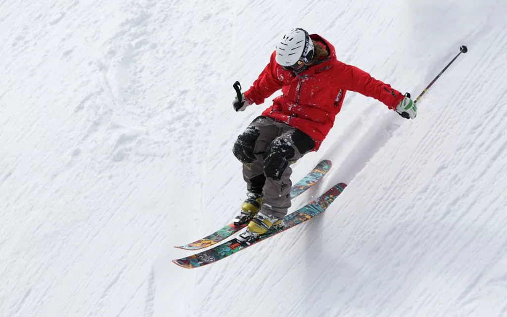 A person in a red jacket is seen skiing fast down the side of a mountain. They have a helmet and goggles, and hold two ski poles.