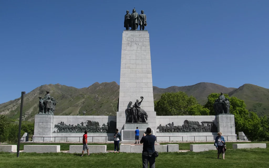 Tall monument on flat grass with Utah mountains behind it. Here you can see the This is The Place Monument located near Salt Lake City.