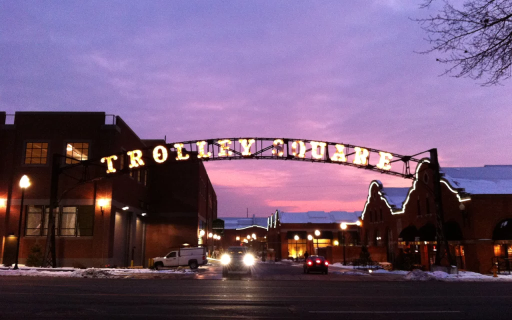 The entrance to Trolley Square in Salt Lake City. The picture is taken at dusk on a snowy evening. One car enters the parking lot, while another leaves.