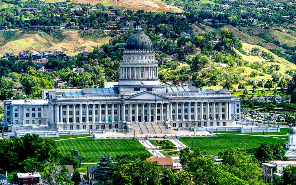 Grand Utah State Capitol Building located in Salt Lake City. This building is reminiscent of the US capital with its columns on the exterior, and dome roof tower. A nicely maintained grassy area sits in front of the building. Behind the building are hills and mountains with houses and buildings.