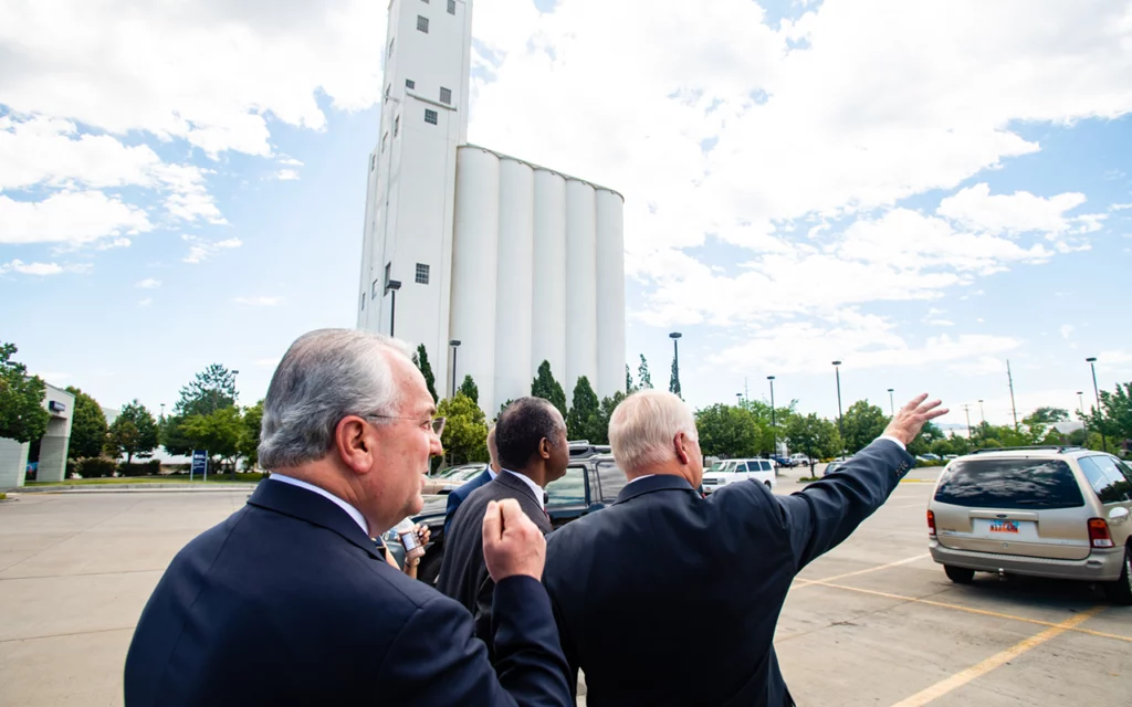 Government officials walk in the parking lot next to Welfare Square in Salt Lake City. One of them points away from the building at something in the distance.