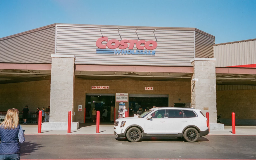 The outside of a Costco Wholesale entrance. One opening in the building says “entrance”. The other says “exit”. Shopper walk up to the store alone, and with carts. There is a white suburban-like car parked in front of the wholesale store.