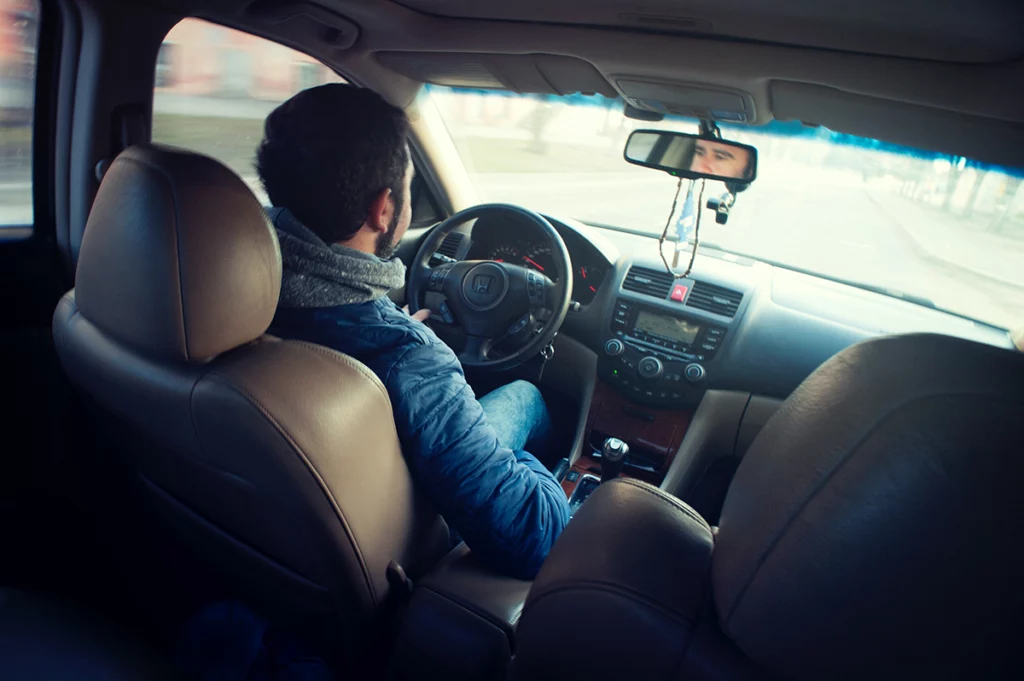 Image shows the interior of a car captured from the backset. The driver is seen wearing denim clothing and has one hand casually on the steering wheel. The interior rear-view mirror shows part of the driver’s face.