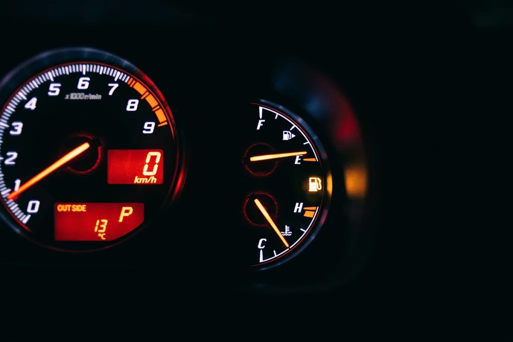 Image shows part of dashboard instrument cluster captured in the dark as numbers, letters, markers, and needles of speedometer, fuel gauge and temperature gauge are lit up. Speedometer needle points at 1, fuel gauge needle hovers at E which indicated empty, and temperature gauge needle is closer to C (cold) than H (hold).