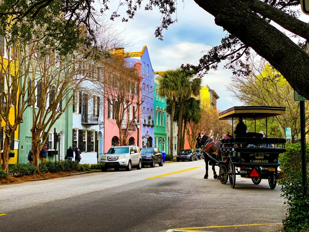 Image shows a row of houses painted in striking hues while there are cars parked on the street and an approaching horse-drawn carriage.